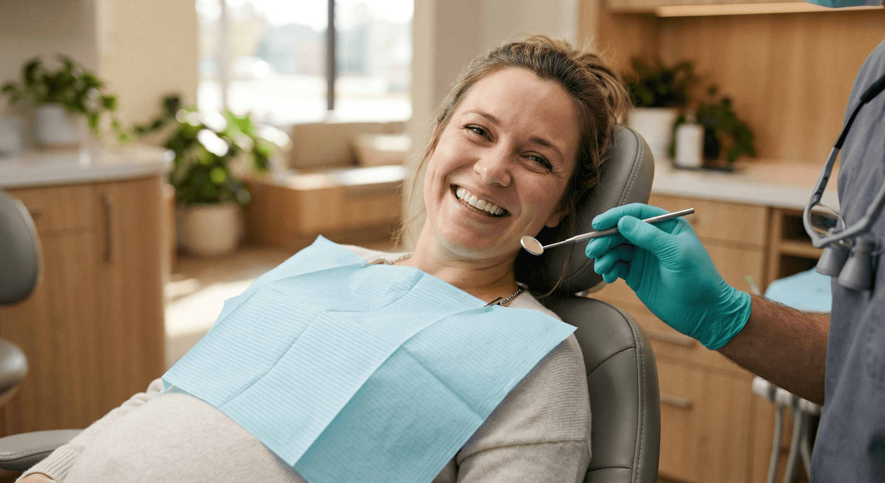 Smiling pregnant woman in dental chair with blue bib, dentist holding mirror nearby, warm modern office with plants
