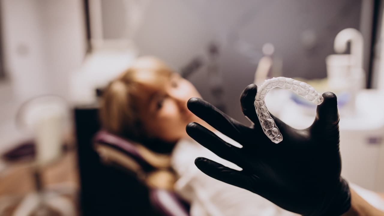 Gloved hand holding a clear Invisalign aligner with a patient reclined in the background