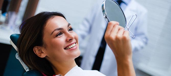 Woman in dental chair smiling at a hand mirror after a cosmetic dental procedure